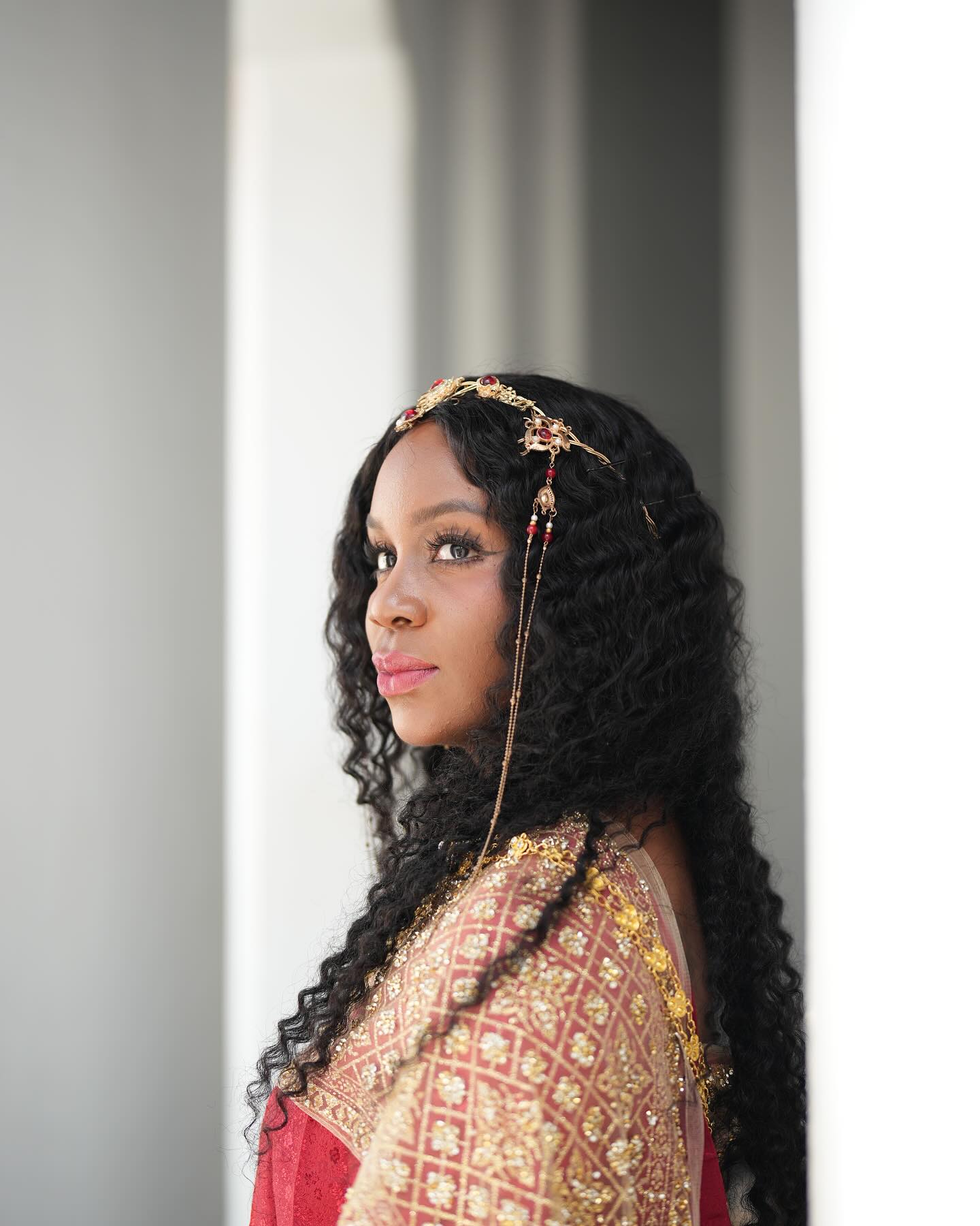 Close portrait of Neké Akua in a red and gold embroidered outfit and a jeweled gold headpiece, looking back over her shoulder against a soft gray stone backdrop.