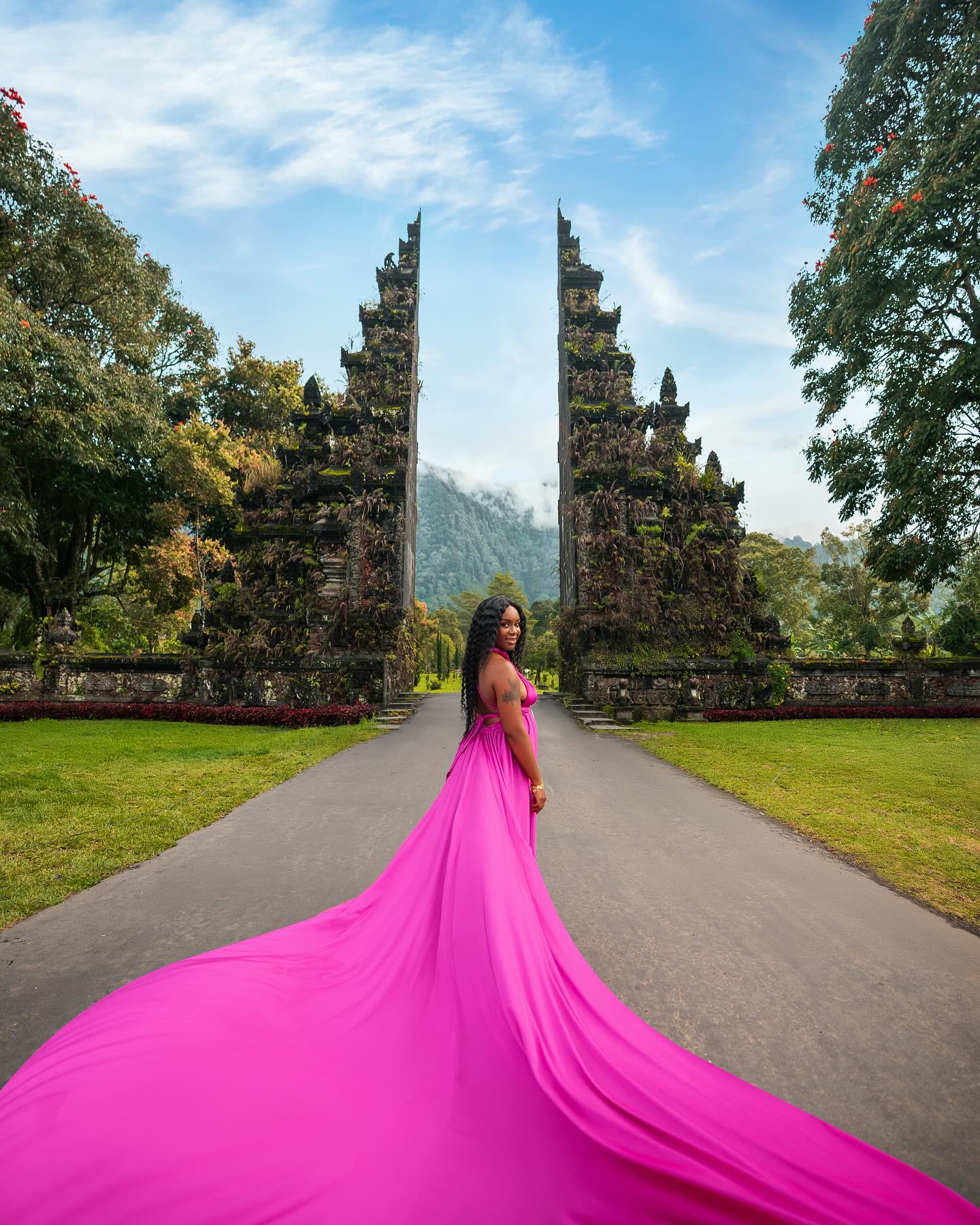 Neké Akua in a magenta gown with a long sweeping train, standing between the two halves of a moss-covered Balinese split gate with jungle mountains behind.