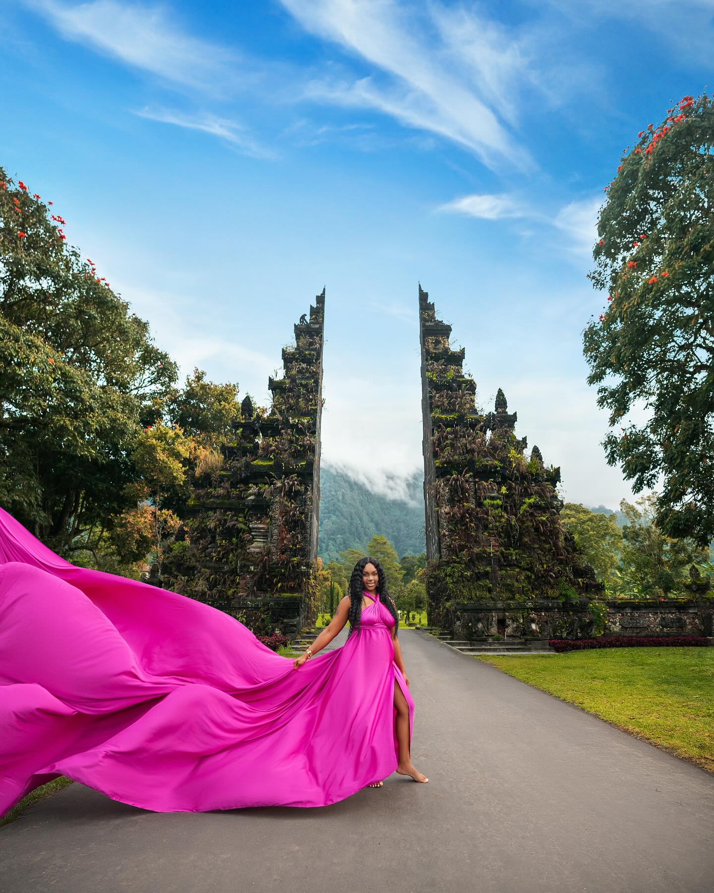 Neké Akua holding out the train of her magenta gown so it sweeps across the road, standing barefoot in front of a Balinese split gate beneath a wide blue sky.