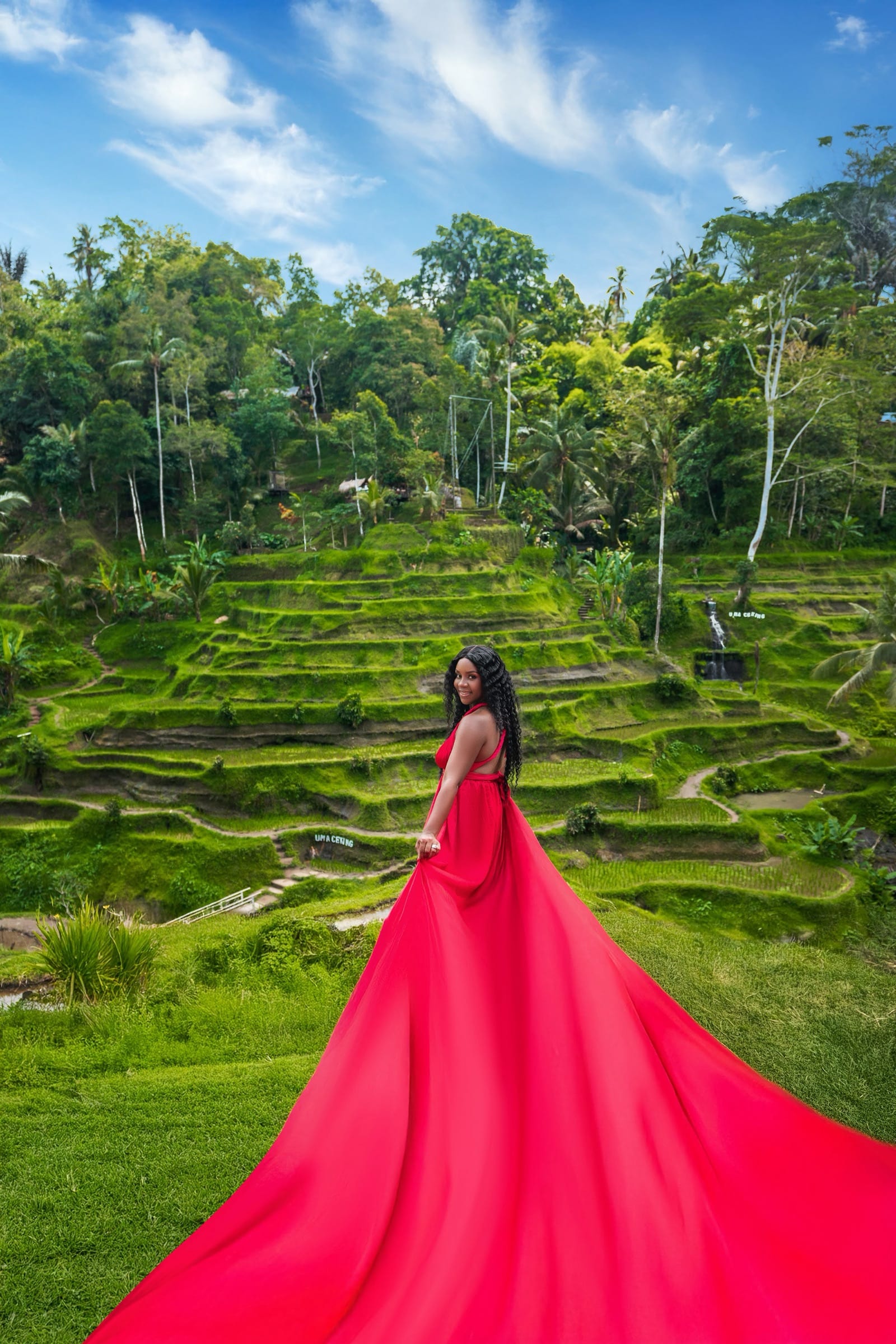 Neké Akua in a flowing red gown on the Tegallalang rice terraces in Bali, looking back over her shoulder as the train of the dress spills down the hillside behind her.