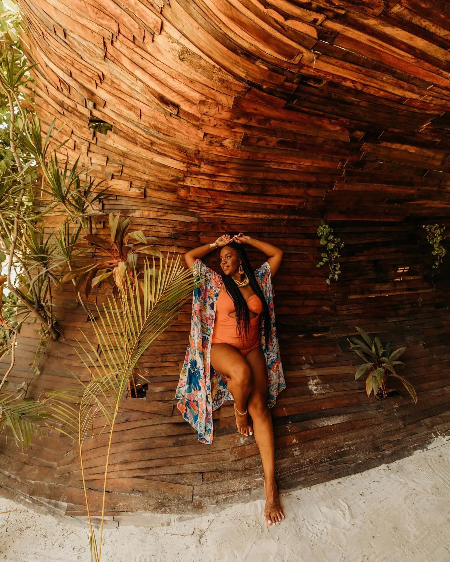 Neké Akua in an orange one-piece swimsuit against a curved wooden cabana wall.